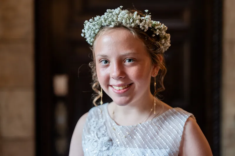 Close-up portrait of a smiling child wearing a white textured sleeveless dress and delicate baby's breath crown in front of stone architecture.