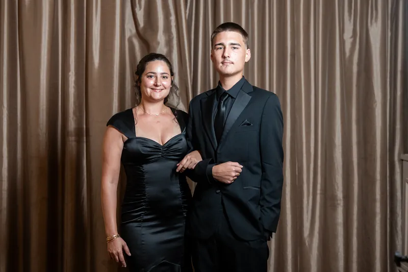 Kimberly and Ty stand arm-in-arm against golden curtains at The Treasury on the Plaza, both in dark formal attire and smiling at the camera.