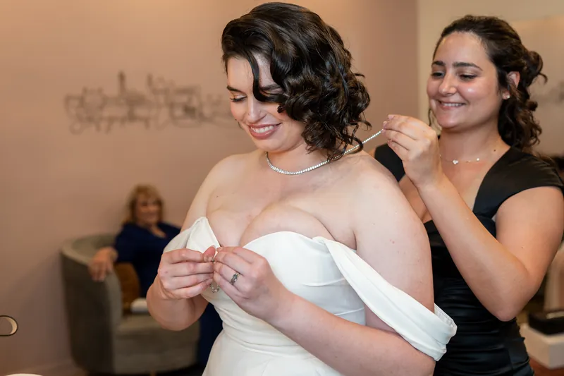 A person in a white strapless gown laughs while another adjusts fabric at her side during bridal preparations.