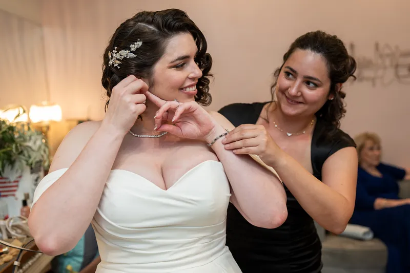 Two people fasten a delicate necklace around the neck of someone wearing an off-shoulder white wedding dress.