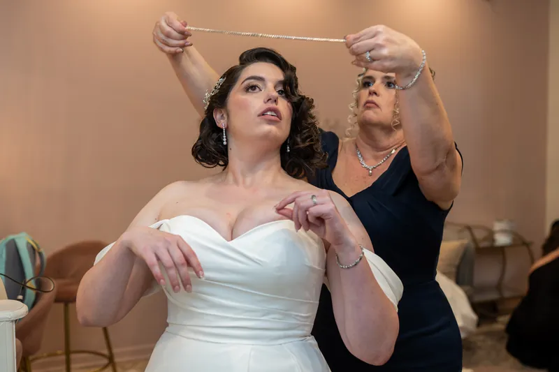 A person in a white gown looks upward while someone behind adjusts a veil with ribbons during getting-ready moments.