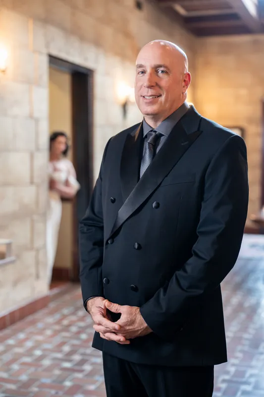 A man in a navy double-breasted suit stands in The Treasury on the Plaza's stone-walled hallway, smiling toward the camera.