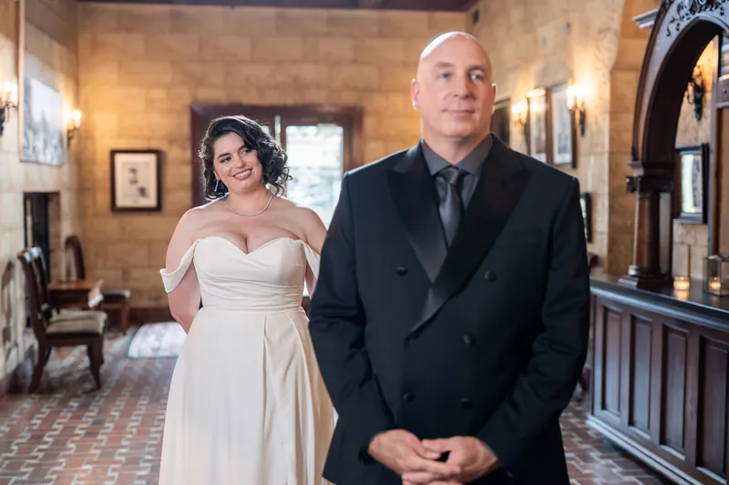 Ty stands in the foreground of The Treasury's elegant corridor in a dark suit while Kimberly waits behind in her white gown.