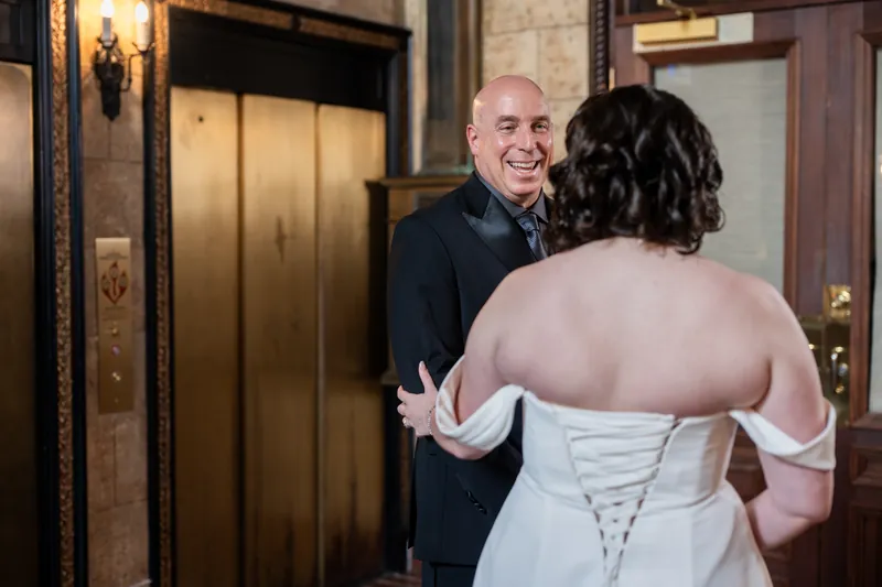 Two people in wedding attire smile at each other in The Treasury's stone lobby near golden elevator doors.
