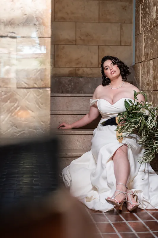 A person in a white off-shoulder gown sits on brick flooring holding a lush green bouquet, gazing downward.