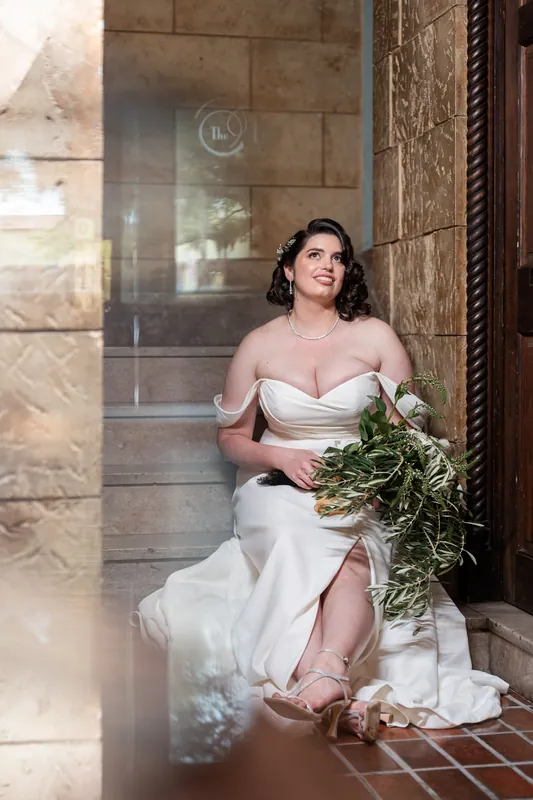 Someone in a flowing white dress cradles a verdant bouquet while seated against textured stone walls at The Treasury.