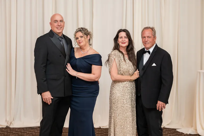 Four family members pose together indoors—two men in dark suits flank two women in formal evening wear against a cream draped backdrop.