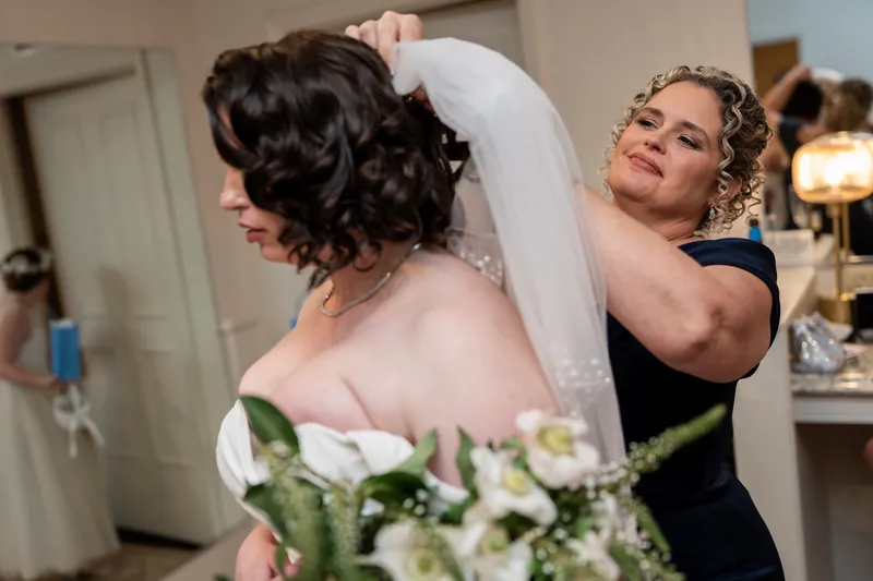 A person in a white wedding gown has their veil adjusted by someone in black during bridal prep.