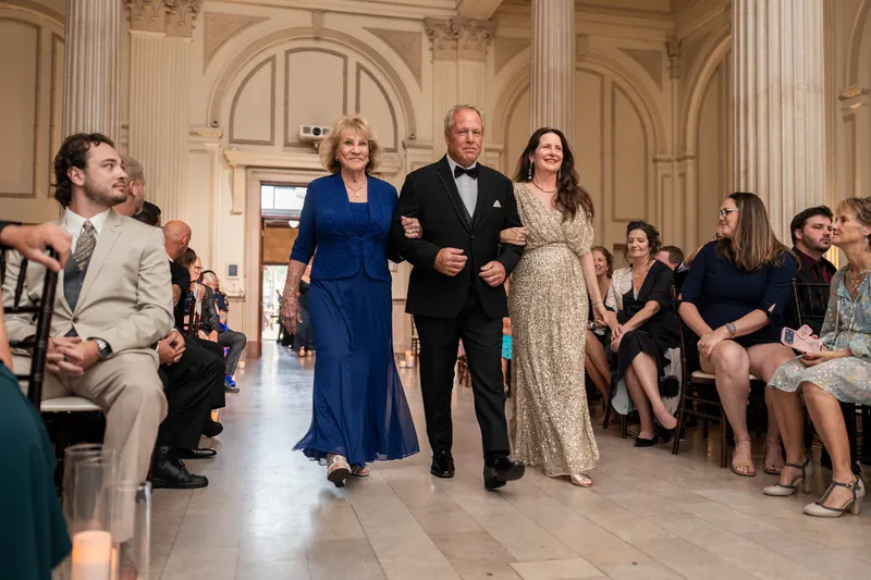 Two people walk down the aisle of The Treasury on the Plaza's ornate hall lined with seated guests in formal attire.
