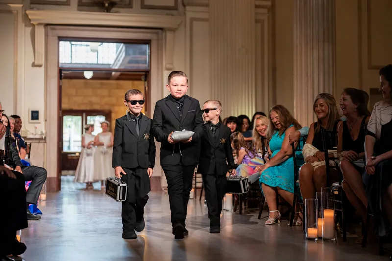 Two young attendants in formal black walk together down the aisle at The Treasury on the Plaza with guests watching from both sides.