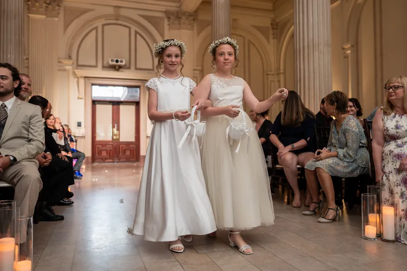 Two people in white gowns stand hand-in-hand in The Treasury's columned hall as seated guests watch from both sides.