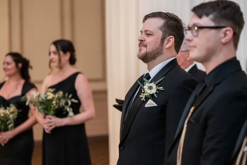 Two men in dark suits stand at the altar, one wearing a yellow boutonniere, watching the ceremony unfold.