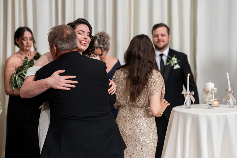 Four adults embrace and laugh together at a reception table, with a fifth person watching in the background.