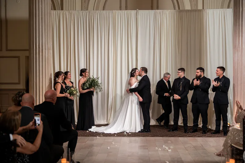 Two people in wedding attire stand at the altar with their wedding party flanking them under draped cream curtains at The Treasury on the Plaza.