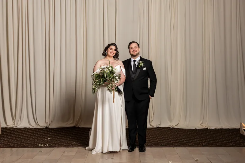 Kimberly and Ty pose together in their wedding clothes, with a cascading greenery bouquet, against pleated ivory backdrop.