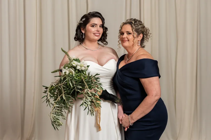 A person in a white gown holds a greenery bouquet while standing beside a woman in a navy off-shoulder dress.