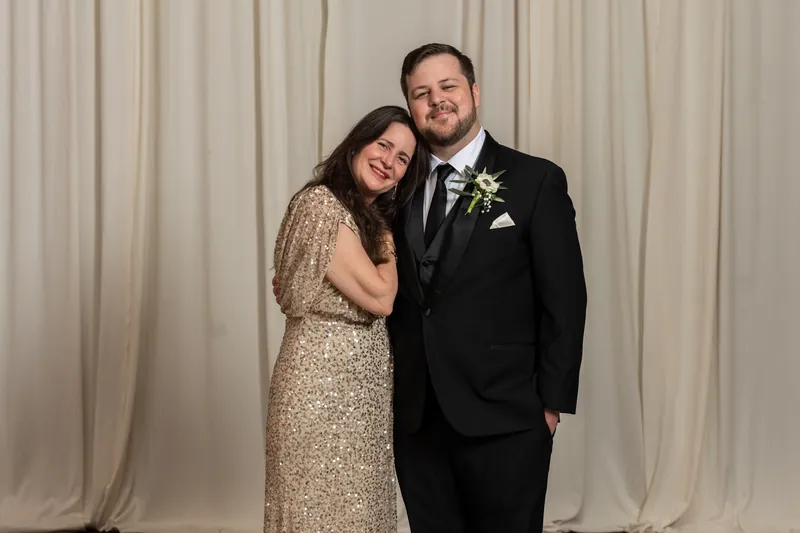 Two people embrace and smile at the camera, with one wearing a gold sequined dress and the other in a black suit with boutonniere.