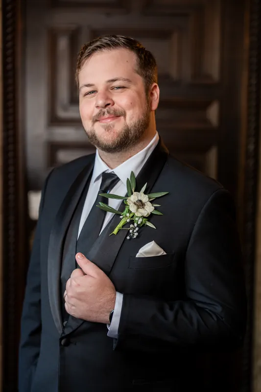 A groom in a dark suit with a white floral boutonniere smiles in a portrait inside the historic venue.