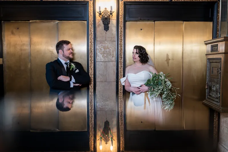 Two people stand on opposite sides of ornate brass elevator doors, each holding greenery and looking toward each other.