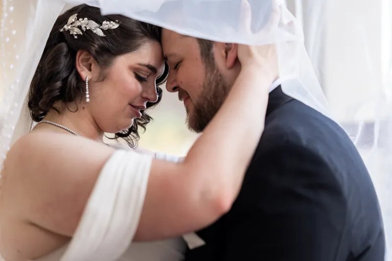 Two people press their foreheads together in an intimate moment, framed by white fabric and natural light.