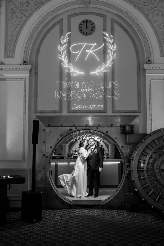 A couple stands beneath an ornate monogrammed arch in a black and white portrait at The Treasury on the Plaza.