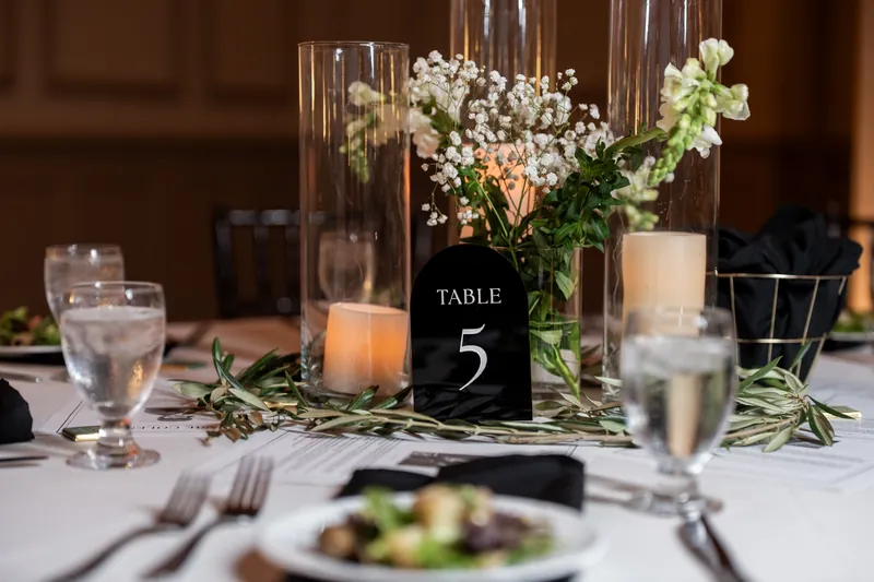Table 5 centerpiece features tall glass cylinders with white baby's breath, green foliage, and candles beside place settings.