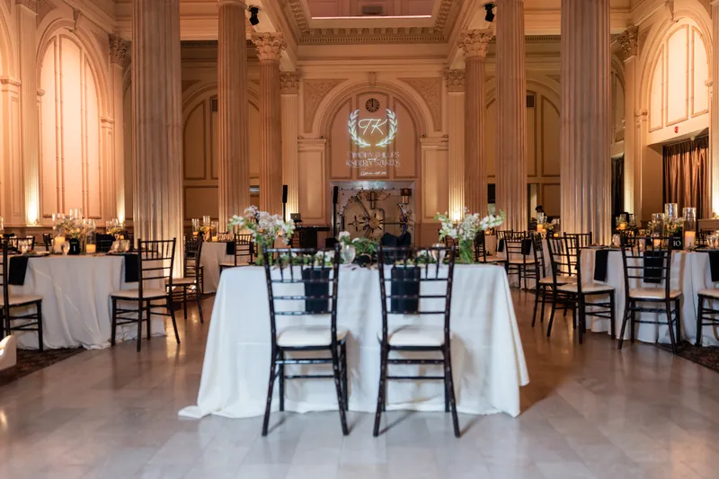 The Treasury on the Plaza's grand hall glows with warm lighting, round tables dressed in white linens arranged throughout the columned space.