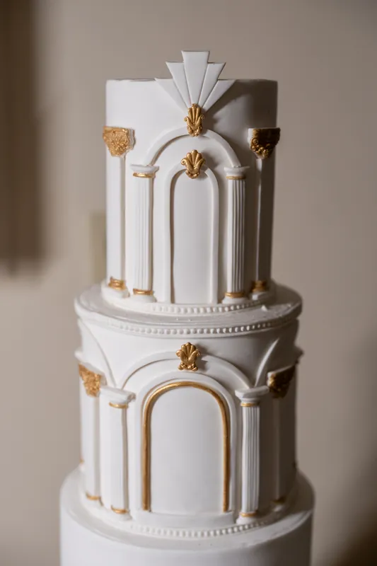 A two-tiered white wedding cake features gold and white art deco arches with matching decorative accents on each tier.