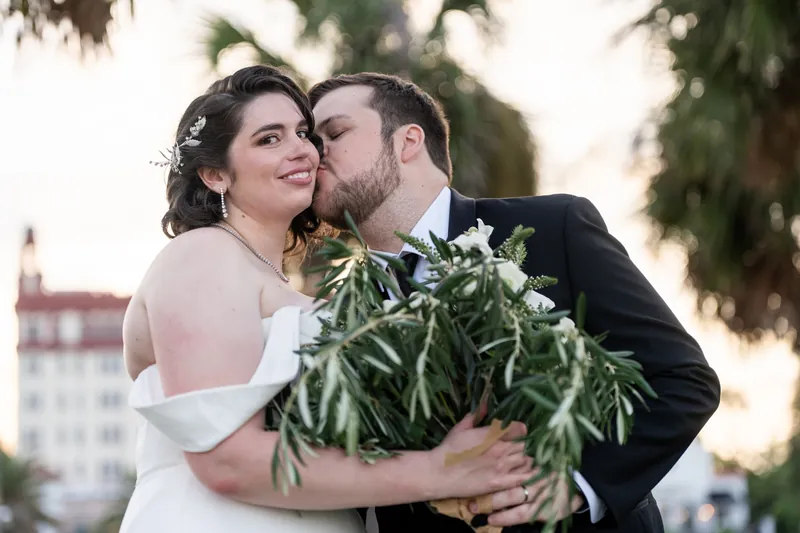 Kimberly and Ty embrace at the bayfront, holding a cascading greenery bouquet as historic buildings frame the background.
