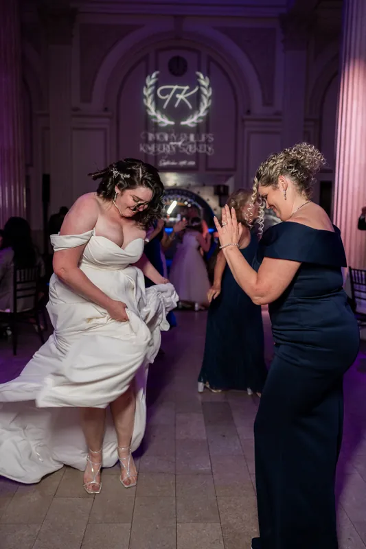 A person in a white gown dances with a woman in black under purple lighting and ornate architectural columns.