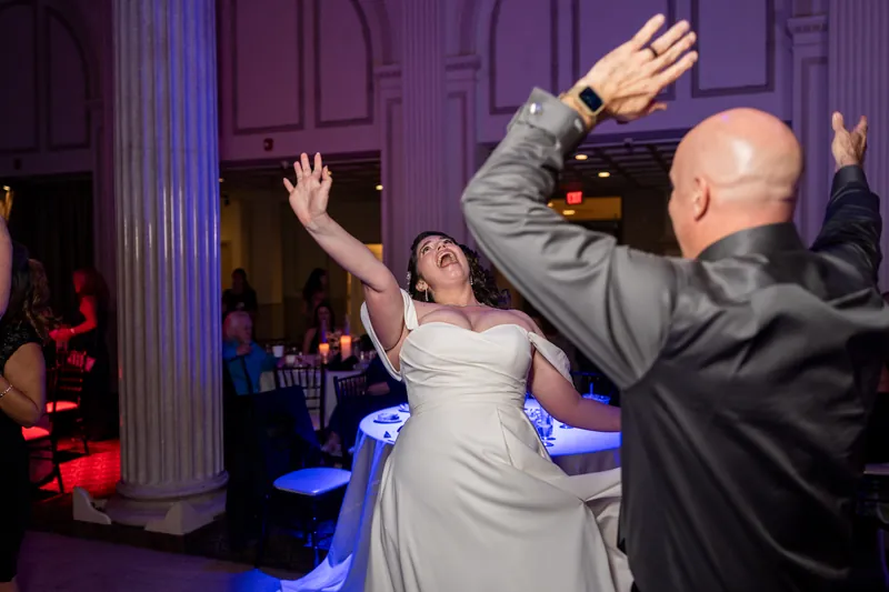 A person in a white gown reaches toward the ceiling while dancing alongside a man in a gray suit and others nearby.