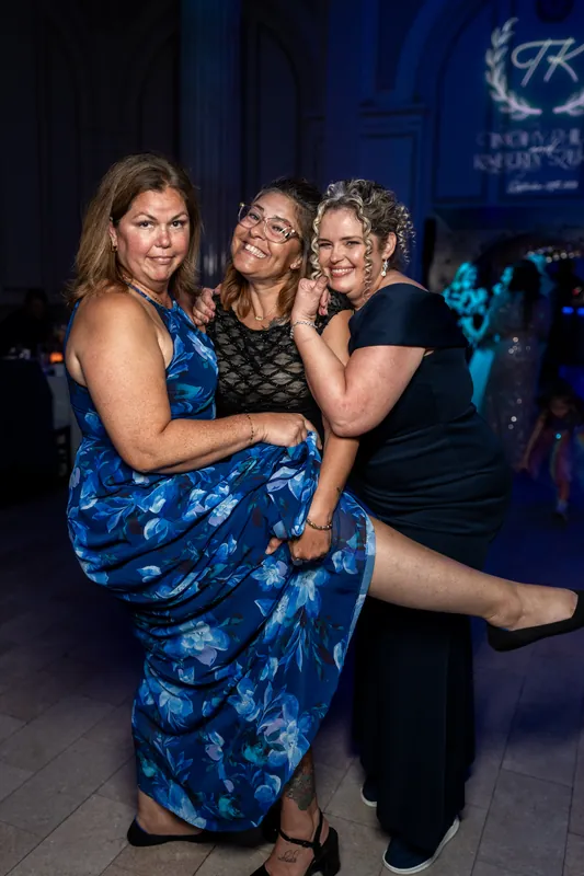 Three women smile and pose together on the dance floor under blue ambient lighting during the reception.
