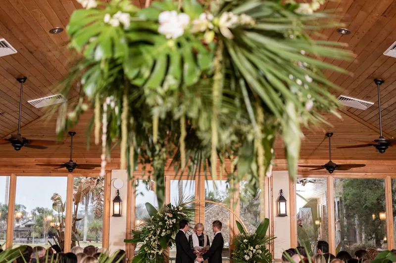 Wide ceremony view from above with hanging tropical greenery and couple at the gold arch