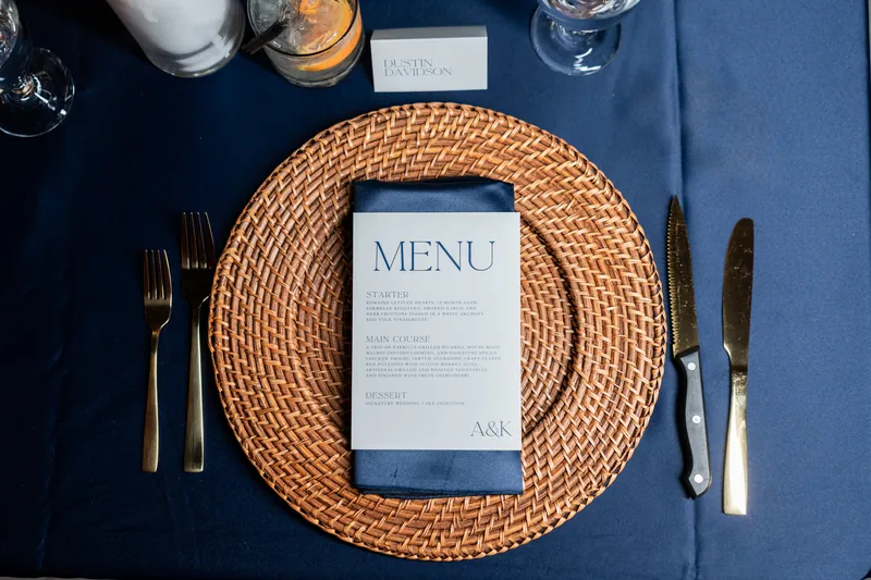 Place setting with navy tablecloth, rattan charger, menu card, and gold flatware