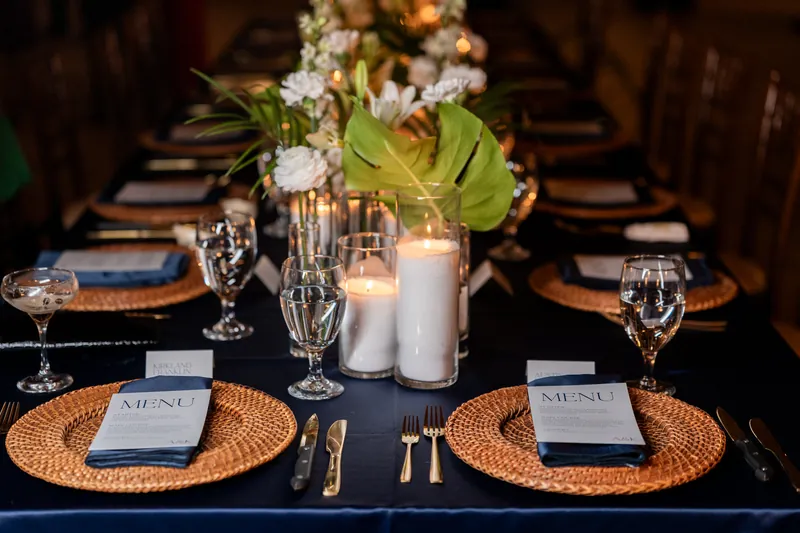 Tablescape with navy linens, tropical centerpiece with monstera and candles, and rattan chargers