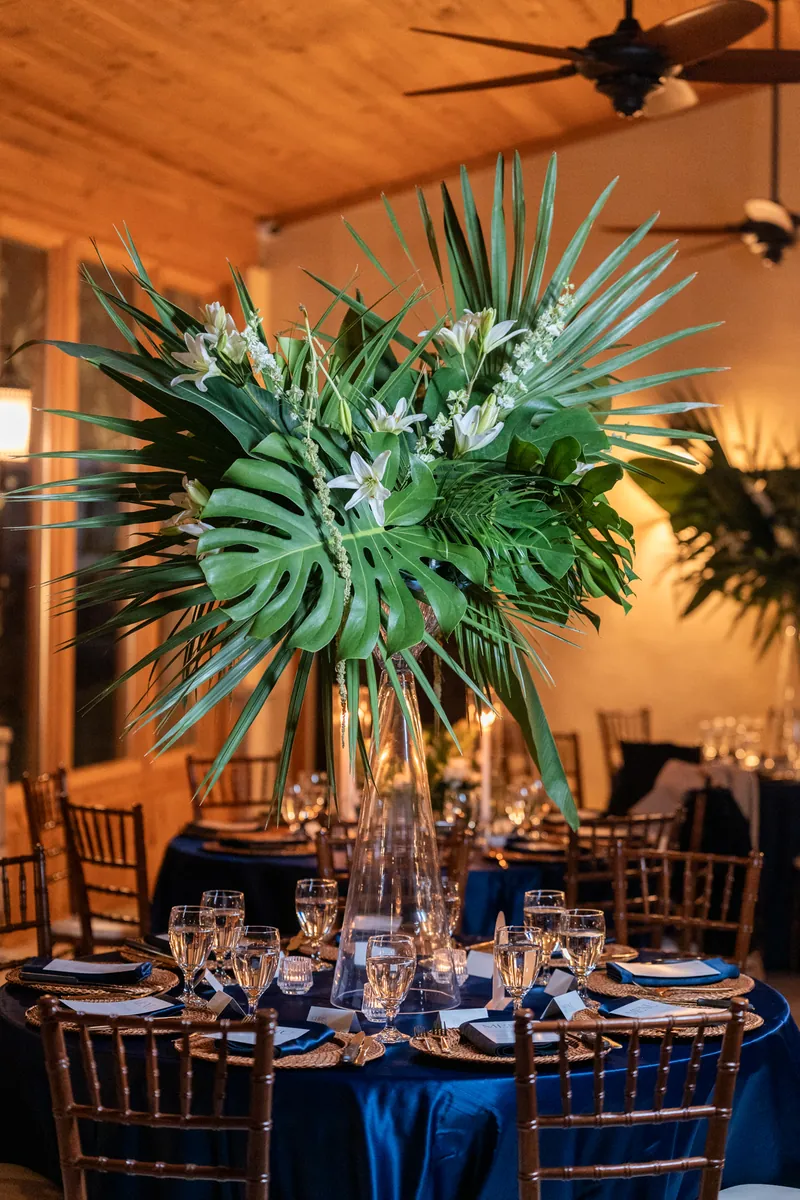 Tall tropical centerpiece with monstera leaves and palm fronds on a round table with navy linens