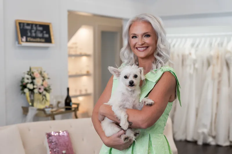 Kristen holds a small white dog while wearing her green dress near white wedding gowns.