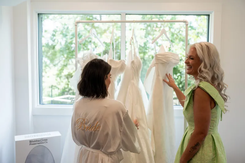 Two women examine a delicate ivory wedding dress hanging by a large window with natural greenery visible outside.