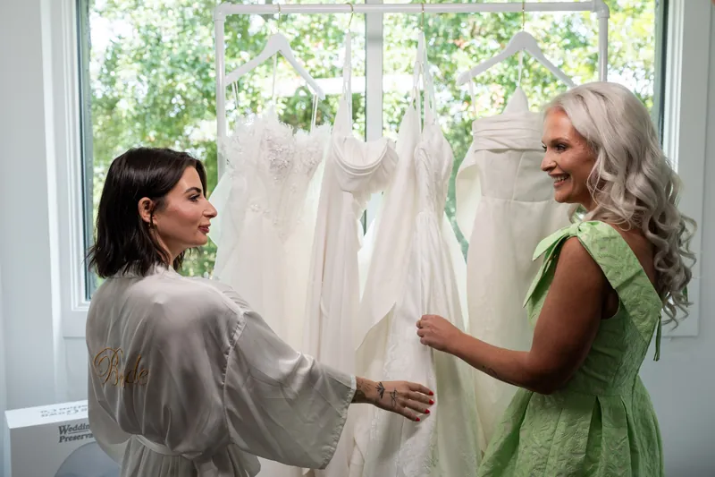 Kristen and a woman discuss an ivory gown displayed between them by a bright window.