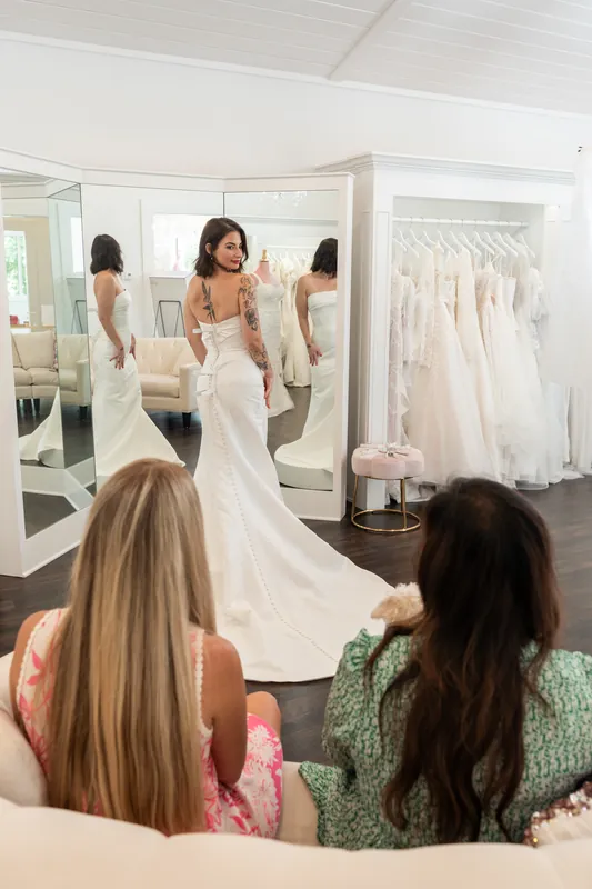 Bridesmaids watch from the foreground as a bride in a flowing white gown poses in the showroom at Tebault Bridal.