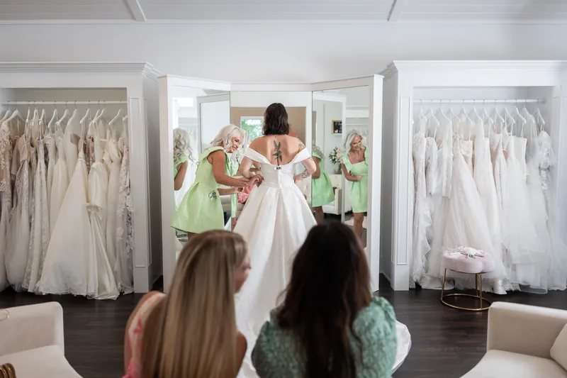 Guests gather around a bride in a white gown at Tebault Bridal, admiring her dress in the mirrored fitting room.