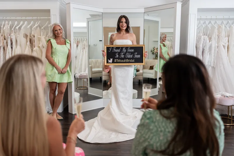 A bride holds a chalkboard sign while modeling her white wedding dress for friends at Tebault Bridal in St. Augustine.