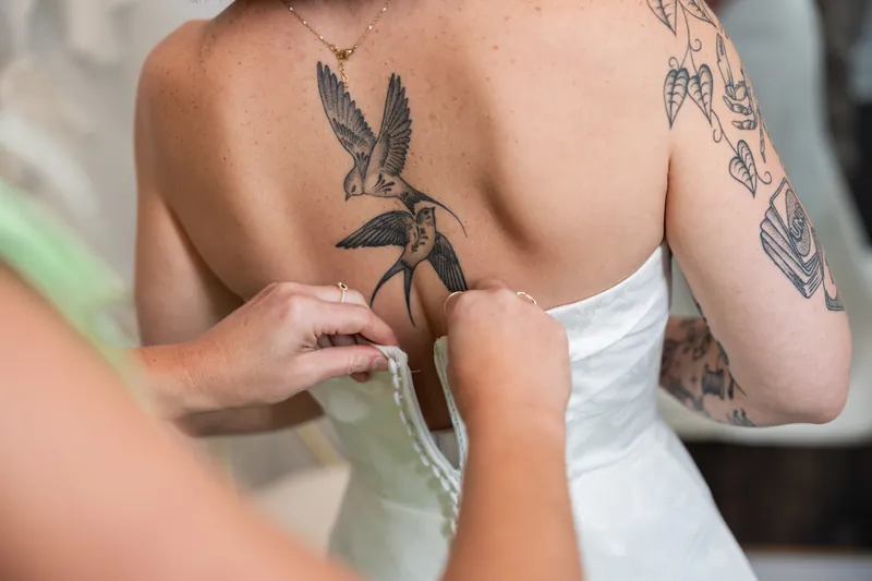 Someone fastens the back of the bride's white gown, revealing her tattooed shoulders and back.