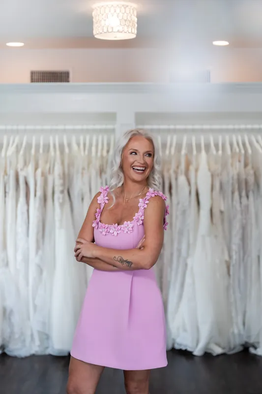 Kristen stands with folded arms between rows of white wedding dresses hanging on either side.
