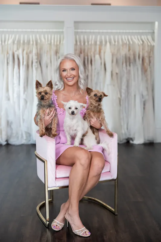 Seated on a pink chair, Kristen holds two small dogs while dressed in her pink outfit at the shop.