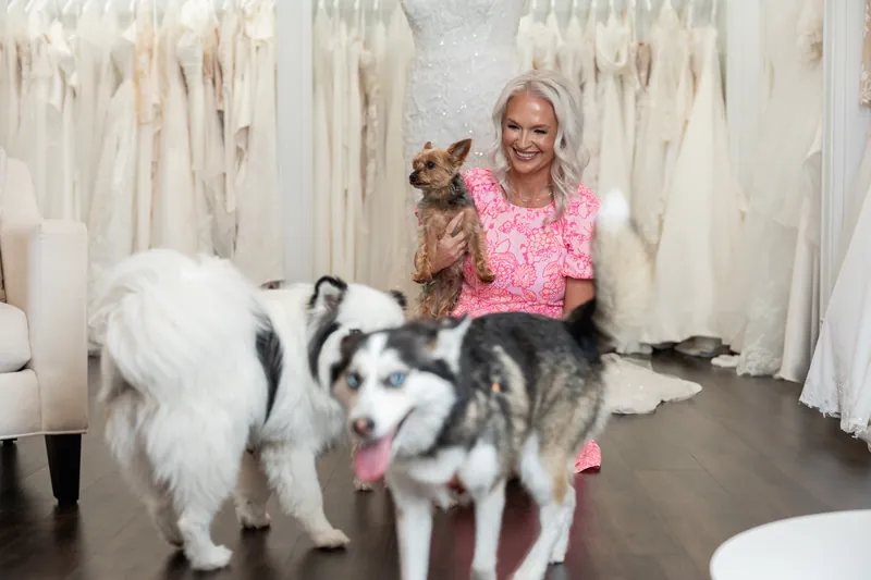 Kristen kneels beside two dogs on the showroom floor, smiling in her pink outfit surrounded by hanging wedding gowns.