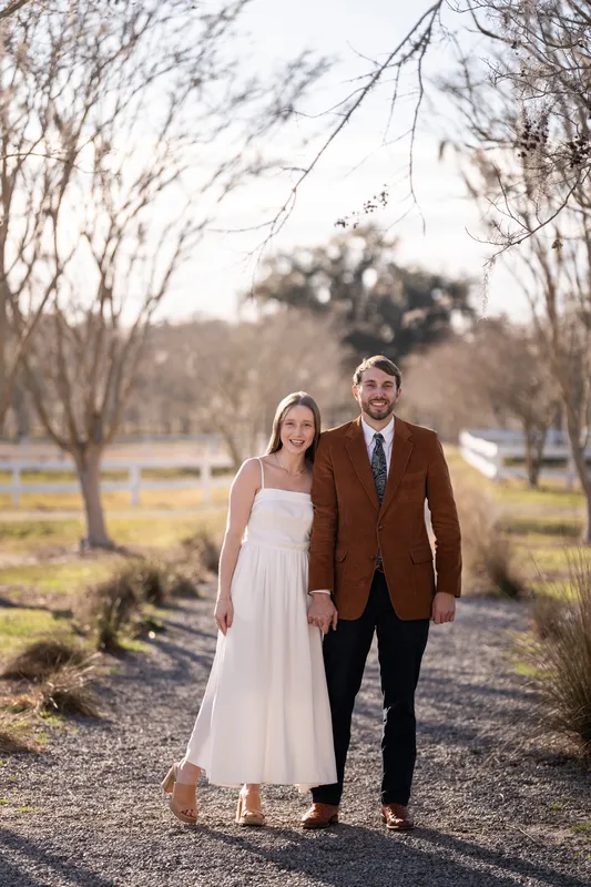 Lacey and Josh standing on a tree-lined gravel path at Congaree and Penn, Lacey in a white dress and Josh in his father's vintage corduroy jacket