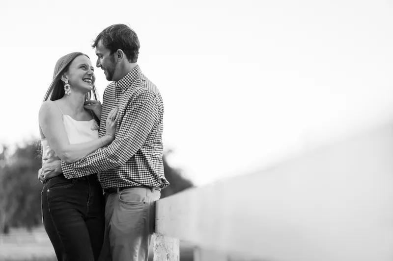 Black and white photo of Lacey laughing while leaning into Josh by a white fence