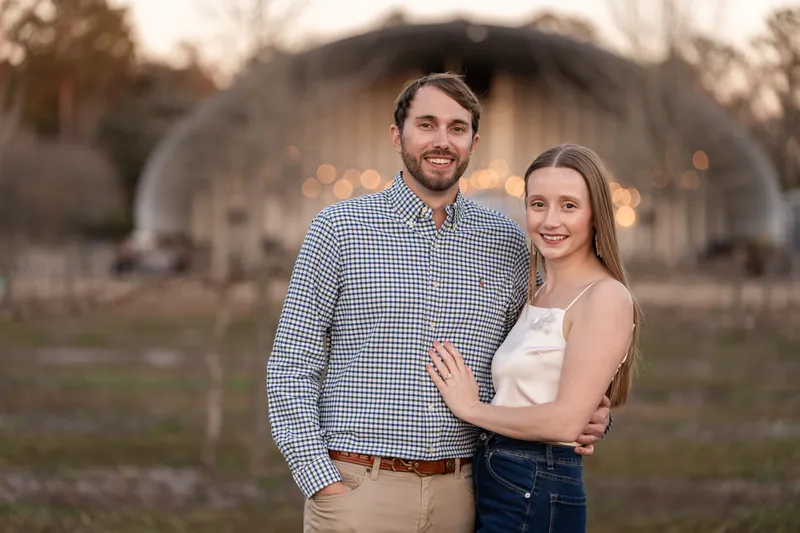 Lacey and Josh in front of the barn with string light bokeh in the background