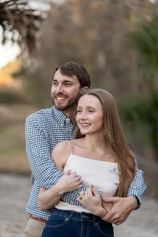 Josh embraces Lacey from behind in a blue plaid shirt while she smiles, with blurred cypress trees and water visible beyond.